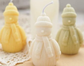 Pode incluir: Três velas em forma de boneco de neve, cada uma com um chapéu e uma echarpe de malha de cor diferente. As velas são brancas, amarelas e verdes.