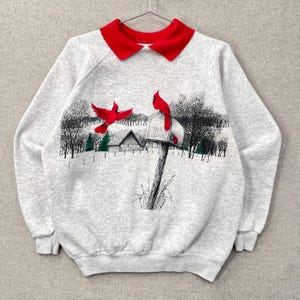 Op de afbeelding: Een grijze sweatshirt met een rode kraag. De voorkant toont een winterse scène met rode kardinaalvogels, een brievenbus en een besneeuwd landschap. Het ontwerp omvat bomen en een huis.