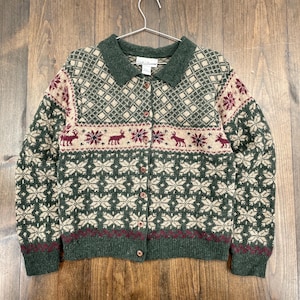 May include: A green, beige, and burgundy button-up cardigan sweater with a collared neckline. The sweater features a snowflake and reindeer pattern. The sweater is hanging on a metal hanger against a wooden background.