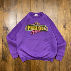 May include: Purple sweatshirt with a vintage Universal Studios Florida logo printed on the front. The logo is in neon colors and features a stylized building with the text "Universal Studios Florida".