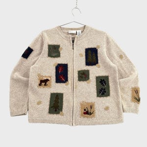 May include: A beige knit zip-up cardigan featuring a variety of rectangular patches with nature-themed embroidery, including a moose, pine trees, oak leaves, and cattails. The garment has a ribbed crew neckline, cuffs, and hem with a central zipper.