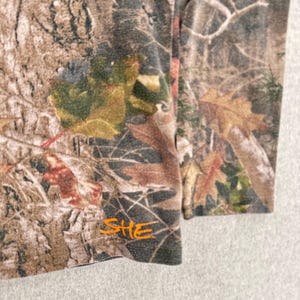 Pode incluir: Uma camisa de manga comprida com um padr&atilde;o de camuflagem florestal realista em tons de castanho, verde e bege de casca de &aacute;rvore e folhas ca&iacute;das. A palavra SHE est&aacute; impressa numa fonte laranja brilhante e estilizada perto da bainha inferior.