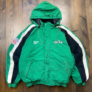 Könnte beinhalten: Grüne Kapuzenjacke mit schwarzen und weißen Akzenten. Mit dem NFL-Logo auf dem linken Ärmel und dem Wort "JETS" auf der Brust. Die Jacke hat ein Reebok-Logo und eine Kapuze mit Kordelzug.
