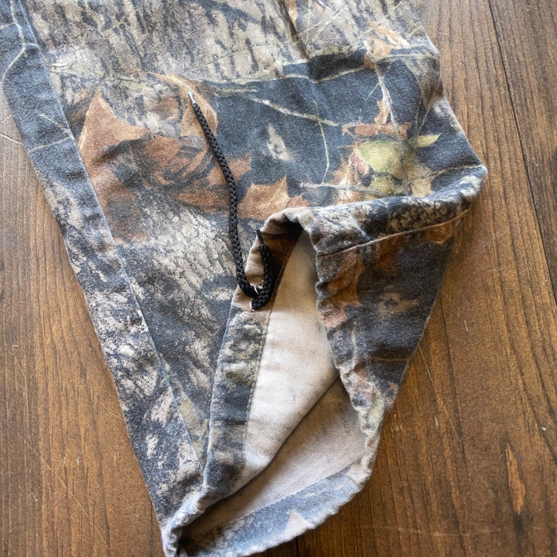 May include: Camouflage fabric with a black drawstring. The fabric is folded in half and the drawstring is visible at the top of the fold.