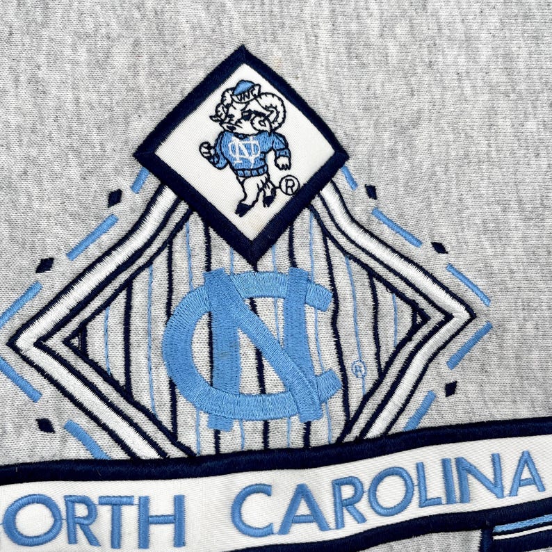 May include: Close-up of a grey sweatshirt with a light blue and white embroidered design. The design includes the words "NORTH CAROLINA" and a cartoon ram mascot. The sweatshirt has a diamond-shaped emblem with the letters "NC".