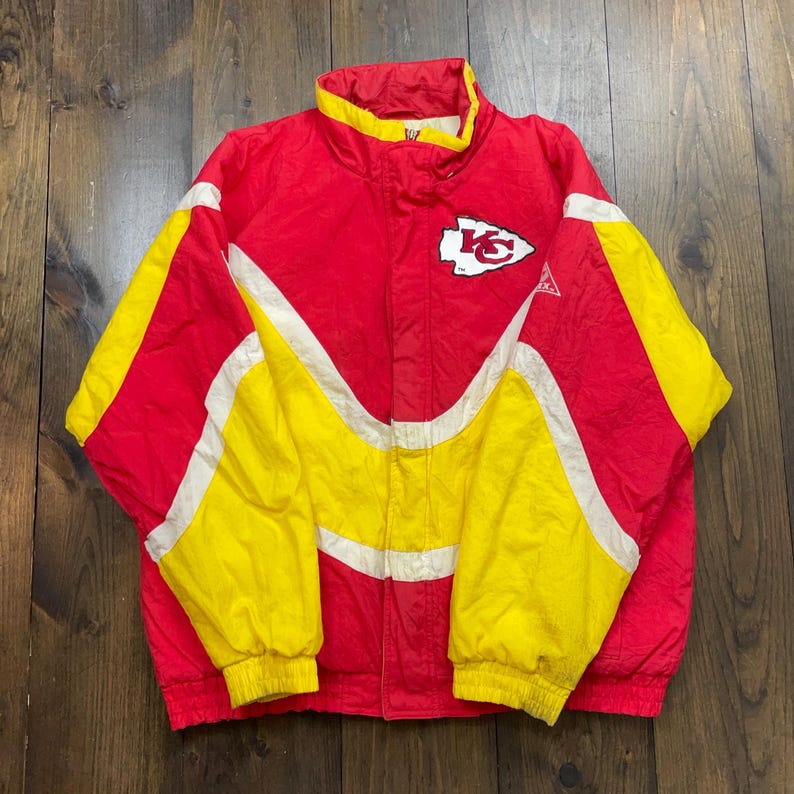 May include: A vintage Kansas City Chiefs jacket in red, white and yellow with a team logo on the front. The jacket has a zip fastening and a stand-up collar.