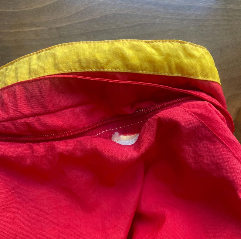 May include: Close-up of a red jacket with a yellow zip and a yellow trim around the collar.