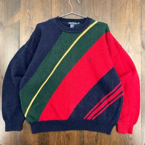 以下が含まれることがあります： ヴィンテージのノーティカセーター。カラーブロックのデザインで、ネイビーブルー、グリーン、レッド、イエローのストライプが特徴です。胸にはノーティカのロゴが見えます。セーターは金属製のハンガーに掛けられています。
