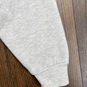 May include: Close-up of a light grey sweatshirt sleeve with a ribbed cuff. The sleeve has several small, light brown stains. The fabric appears to be a soft, knit material. The background is a wooden surface.