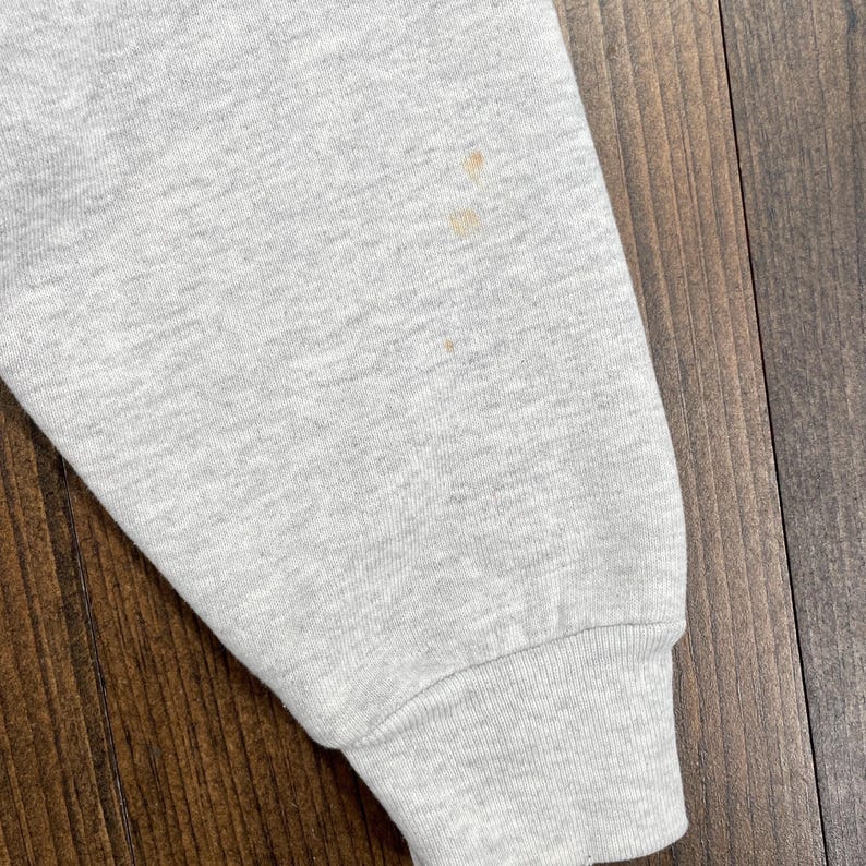 May include: Close-up of a light grey sweatshirt sleeve with a ribbed cuff. The sleeve has several small, light brown stains. The fabric appears to be a soft, knit material. The background is a wooden surface.