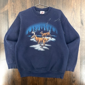May include: Navy blue sweatshirt with a graphic of two deer running through a snowy landscape with a forest backdrop. The deer are brown with white accents, and the forest is a dark blue. The sweatshirt has a crew neck and long sleeves.