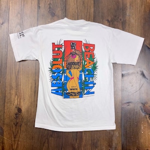 Peut inclure: T-shirt blanc avec un graphisme coloré. Le motif présente les mots "Absolut Beach Babe" et une illustration stylisée d'une femme en maillot de bain. Le design comprend des feuilles de palmier et le mot "Imported".