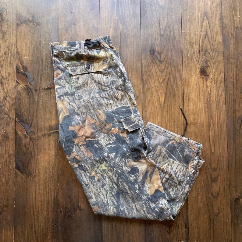 May include: Camouflage cargo pants with a drawstring at the bottom. The pants are made of a durable fabric and have multiple pockets.