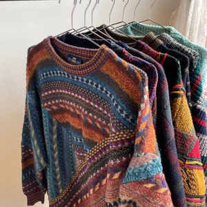 May include: A collection of long-sleeved knit jumpers hanging on a metal rack. Each jumper features a complex, multi-coloured geometric pattern with various textures, including shades of blue, red, yellow, green, and brown in intricate designs.