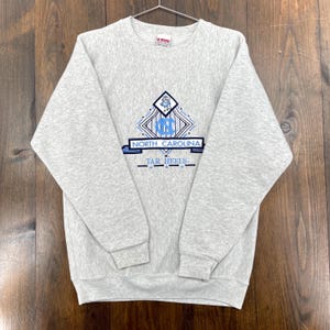 May include: A grey sweatshirt featuring a North Carolina Tar Heels design. The design includes a light blue diamond shape with "NC", and the words "North Carolina" and "Tar Heels" in blue.