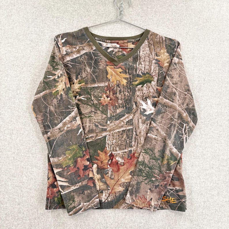Pode incluir: Uma camisola de manga comprida com decote em V, apresentando um padr&atilde;o de camuflagem florestal realista com texturas de casca de &aacute;rvore castanha e folhas de outono espalhadas em tons de verde, amarelo e laranja. O decote tem um rebordo verde-azeitona.