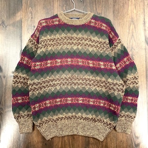 May include: A vintage crew neck sweater with a colorful geometric pattern. The sweater features a mix of green, purple, red, and tan colors. The knitwear is hanging on a metal hanger. The sweater has a ribbed collar, cuffs, and hem.