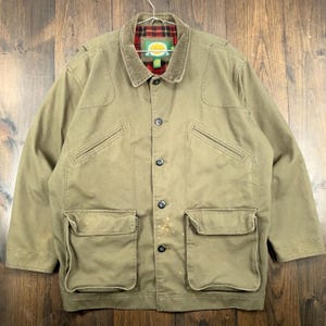 Puede incluir: Chaqueta de color caqui con cuello de pana y forro a cuadros rojos y negros. La chaqueta tiene cuatro bolsillos delanteros, dos bolsillos en el pecho y cierre de botones. El logotipo de Cabela's es visible en el cuello.