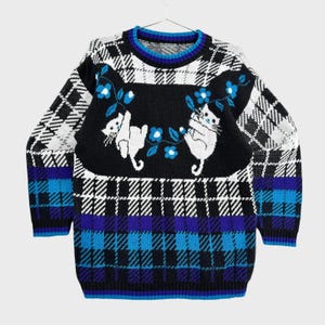 Puede incluir: Un suéter de punto de manga larga con un panel central negro que muestra dos gatos blancos jugando con flores azules. El suéter presenta un patrón de cuadros en negro, blanco y tonos azules, con cuello redondo y puños acanalados.