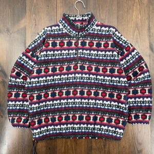 May include: A patterned fleece pullover with a half-zip. The design features horizontal bands of red, white, black, and blue geometric shapes. The fleece is hanging on a metal hanger against a wooden background.