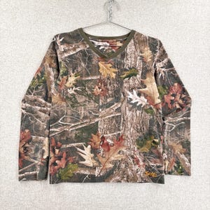 Pode incluir: Uma camisa de manga comprida com decote em V apresentando um padr&atilde;o de camuflagem florestal realista com folhas de carvalho e texturas de casca de &aacute;rvore em tons de castanho, verde e bege. Possui um acabamento verde-oliva no decote e a palavra SHE bordada em laranja.