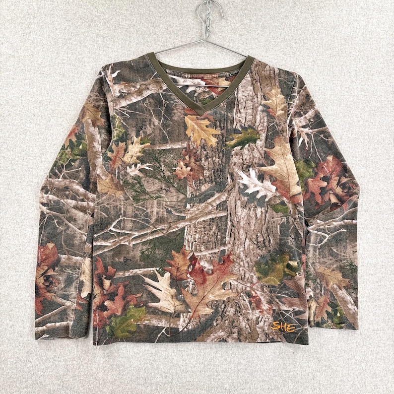 Pode incluir: Uma camisa de manga comprida com decote em V apresentando um padr&atilde;o de camuflagem florestal realista com folhas de carvalho e texturas de casca de &aacute;rvore em tons de castanho, verde e bege. Possui um acabamento verde-oliva no decote e a palavra SHE bordada em laranja.