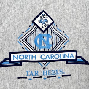 May include: A grey sweatshirt with the North Carolina Tar Heels logo. The design features the words "North Carolina" and "Tar Heels" in blue and white, a diamond-shaped emblem, and a cartoon mascot.