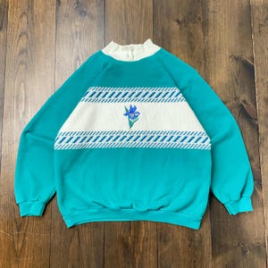 Puede incluir: Sudadera vintage turquesa y blanca con cuello alzado y manga larga. La parte delantera presenta una franja horizontal blanca con detalles geométricos azules y blancos y un bordado floral azul. La sudadera es de material suave.
