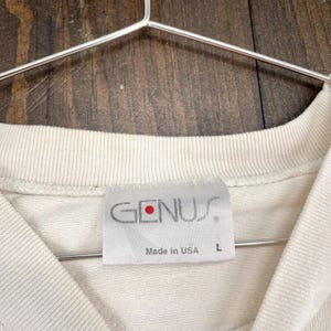 May include: Close-up of a cream-colored t-shirt with a tag that reads "GENUS" in gray lettering with a red dot. The tag also says "Made in USA" and has a size "L". The shirt is hanging on a metal hanger.