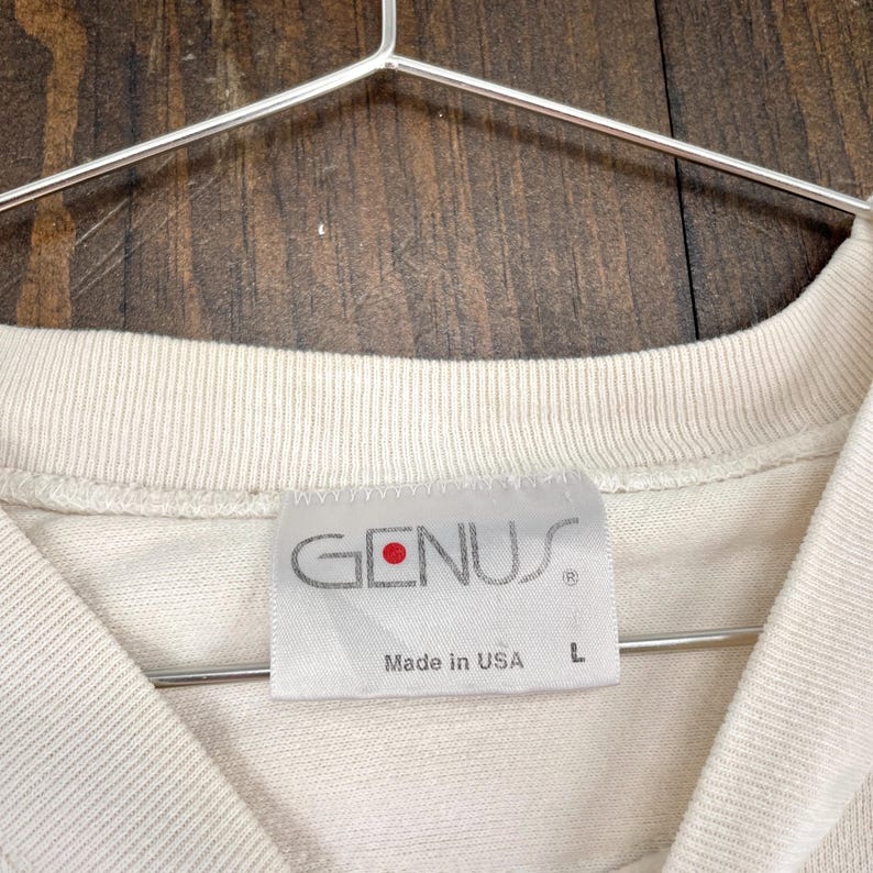 May include: Close-up of a cream-colored t-shirt with a tag that reads "GENUS" in gray lettering with a red dot. The tag also says "Made in USA" and has a size "L". The shirt is hanging on a metal hanger.
