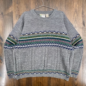 Op de afbeelding: Een grijze trui met ronde hals en een patroon over de borst en mouwen. Het patroon bevat banden in wit, groen, blauw en kastanjebruin. De trui hangt aan een metalen hanger tegen een houten achtergrond.