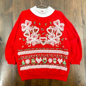 Könnte beinhalten: Ein roter Weihnachtspullover mit weißem Rollkragen. Der Pullover hat weiße Schleifen, Herzen und festliche Verzierungen. Eine weiße Spitzentrimmung verläuft über den unteren Rand. Der Pullover hängt an einem Metallbügel.
