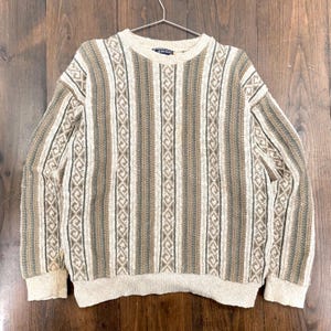 May include: Beige, brown, and green striped sweater with a crew neck. The sweater features vertical stripes with a diamond pattern. The long-sleeved sweater is hanging on a metal hanger against a wooden background.