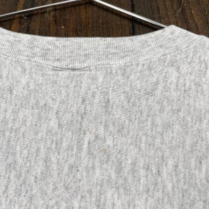 May include: Close-up of a grey crew neck sweatshirt. The collar is ribbed and the fabric has a textured appearance. The sweatshirt is hanging on a metal hanger against a wooden background.