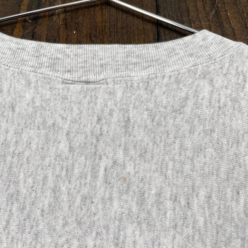 May include: Close-up of a grey crew neck sweatshirt. The collar is ribbed and the fabric has a textured appearance. The sweatshirt is hanging on a metal hanger against a wooden background.