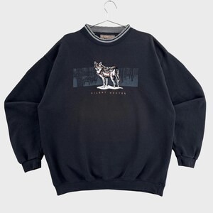 Peut inclure: Un sweat-shirt noir à manches longues orné d'un motif représentant un loup dans une forêt, avec l'inscription "SILENT HUNTER" en dessous. Le vêtement présente un col rond côtelé avec un détail gris à l'intérieur et des poignets côtelés.