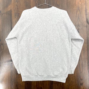 May include: A heather grey sweatshirt hangs on a metal hanger. The crew neck sweatshirt is made of a soft, textured material. The long-sleeved top has a ribbed hem and cuffs. The background is a wooden surface.
