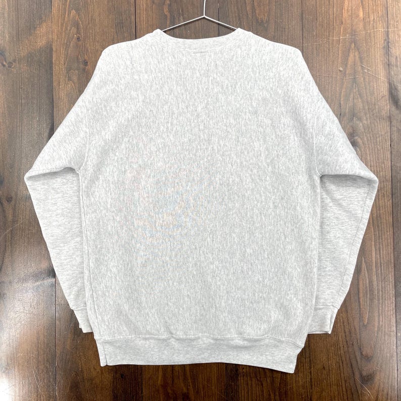 May include: A heather grey sweatshirt hangs on a metal hanger. The crew neck sweatshirt is made of a soft, textured material. The long-sleeved top has a ribbed hem and cuffs. The background is a wooden surface.