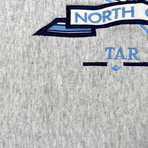 May include: Close-up of grey fabric with embroidered text and design. The word "NORTH" is in blue letters on a white banner. Below, the word "TAR" is embroidered in blue, with a blue diamond shape.
