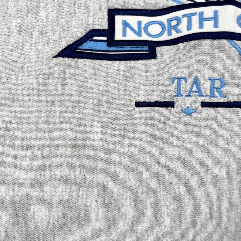 May include: Close-up of grey fabric with embroidered text and design. The word "NORTH" is in blue letters on a white banner. Below, the word "TAR" is embroidered in blue, with a blue diamond shape.