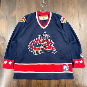 Puede incluir: Jersey de hockey azul marino con detalles rojos y blancos. Presenta un gran logotipo en la parte delantera con una estrella y las letras "CB". El jersey tiene rayas rojas en las mangas y la parte inferior, con estrellas blancas y un logotipo en las mangas.