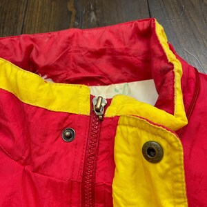 May include: Red and yellow jacket with a zip and two metal buttons. The jacket has a high collar.