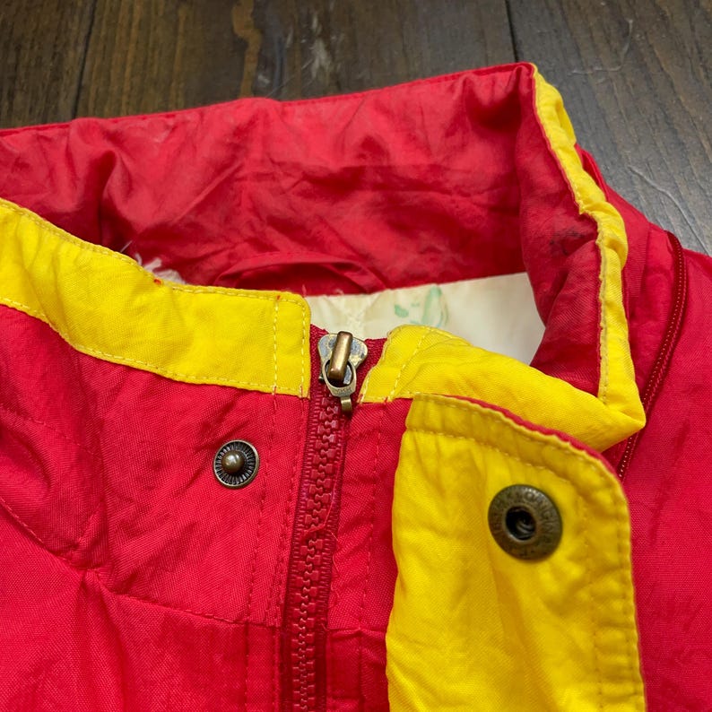 May include: Red and yellow jacket with a zip and two metal buttons. The jacket has a high collar.