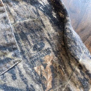 May include: Close-up of a camouflage fabric with a small logo that reads "MOUNTAIN". The fabric is a mix of brown, green, and black.
