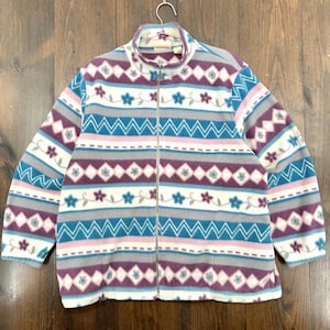 Puede incluir: Chaqueta polar con cremallera completa con un estampado floral y geométrico en tonos azules, morados, grises y blancos. La chaqueta tiene un cuello alto y está colgada de una percha metálica.