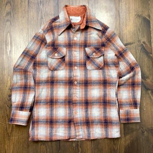 May include: A vintage button-down shirt with a plaid pattern in orange, navy blue, and light blue. The shirt has two chest pockets and a collared neckline. The interior collar is a solid orange color.
