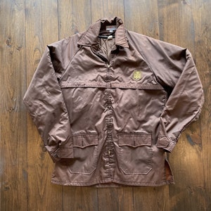 Puede incluir: Chaqueta marrón de UPS con cuello, cierre de cremallera y dos bolsillos delanteros. El logotipo de UPS está en la parte superior derecha del pecho. La chaqueta tiene un acabado satinado y está sobre una superficie de madera.