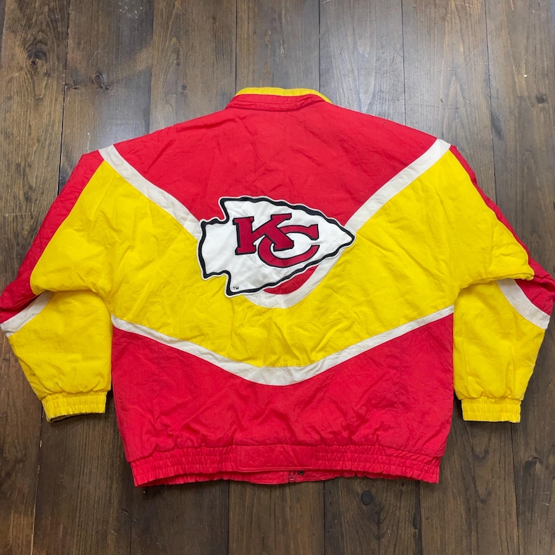 May include: A vintage Kansas City Chiefs jacket in red and yellow with a white and red team logo on the back. The jacket has a zip fastening and a white stripe down each sleeve.