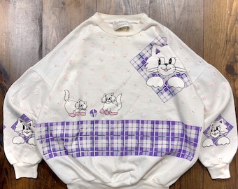May include: White sweatshirt with a purple and white plaid pattern and a cartoon cat design. The sweatshirt has a small pink floral pattern throughout.