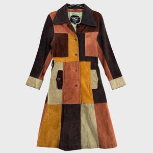 Può includere: Un cappotto a maniche lunghe realizzato con pannelli in pelle scamosciata patchwork in varie tonalità di marrone, marrone chiaro e verde oliva. Il capo presenta un colletto marrone scuro, chiusura frontale con bottoni e due tasche laterali.